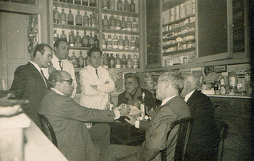 Tertúlia a la rebotiga, els anys cinquanta. D'esquerra a dreta, el fotògraf M. Coria, J. Fluvià, Enrique, Joaquin, el pintor Mercader, Roure i l'arquitecte Sixte. Fotografia de la farmàcia Caldés.
